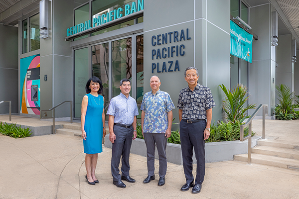 Four people stand outside Central Pacific Plaza in front of a Central Pacific Bank branch entrance, posing together near the building's signage and landscaping. The modern building façade and bank branding are prominently displayed in the background.