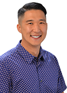 A professional headshot of a person wearing a blue patterned button-down shirt, photographed against a light gray background.