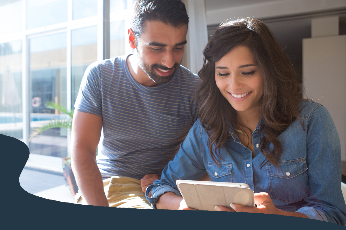 Two people sit together indoors looking at a tablet, with sunlight streaming through large windows in the background. The image highlights digital banking or financial planning, showing users reviewing information on a mobile device in a bright home setting.