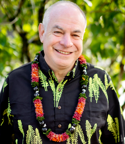 A professional outdoor portrait of a person wearing a black aloha shirt with a green fern pattern and a flower lei, standing against a background of lush green foliage.