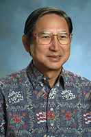 A professional headshot of a person wearing glasses and a multicolored patterned aloha shirt, photographed against a blue studio background.