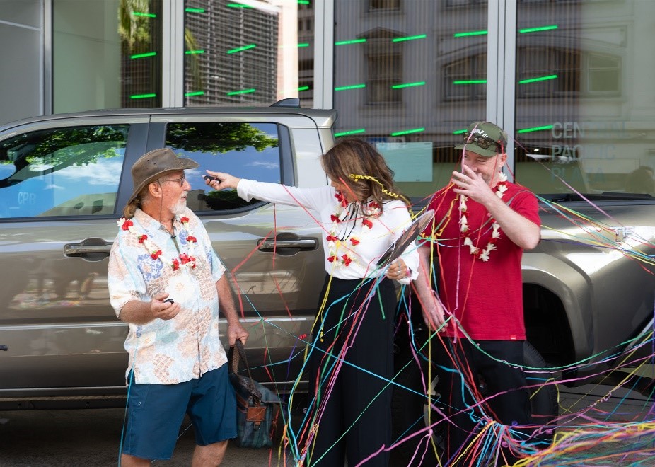A person and two event hosts stand in front of a Toyota Tacoma during a vehicle giveaway celebration. Colorful streamers fill the scene as the hosts present the prize and the truck is displayed in the background.