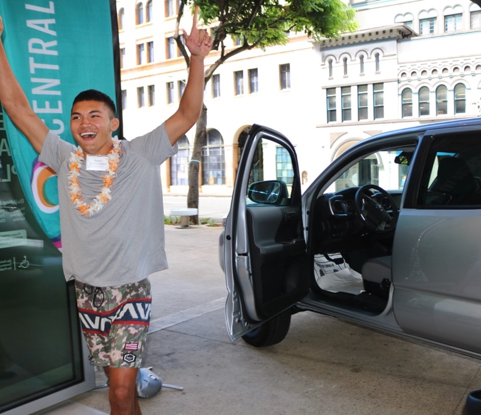 A person wearing a flower lei stands beside an open-door silver Toyota Tacoma outside a Central Pacific Bank branch, raising both arms during a vehicle giveaway celebration. Downtown buildings and Central Pacific Bank branding are visible in the background.