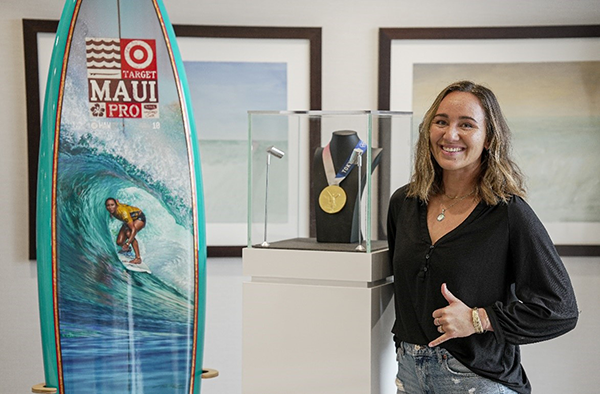 A person stands beside a display case holding a gold medal, next to a surfboard featuring artwork of a surfer riding a large wave. Framed ocean photographs hang in the background, highlighting a surfing-related achievement or exhibit.