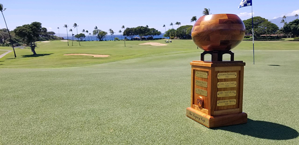 A wooden trophy stands on a golf course overlooking the ocean, with palm trees, putting greens, and sand bunkers visible in the background. The trophy features engraved plaques and sits on the neatly maintained green under a clear blue sky.