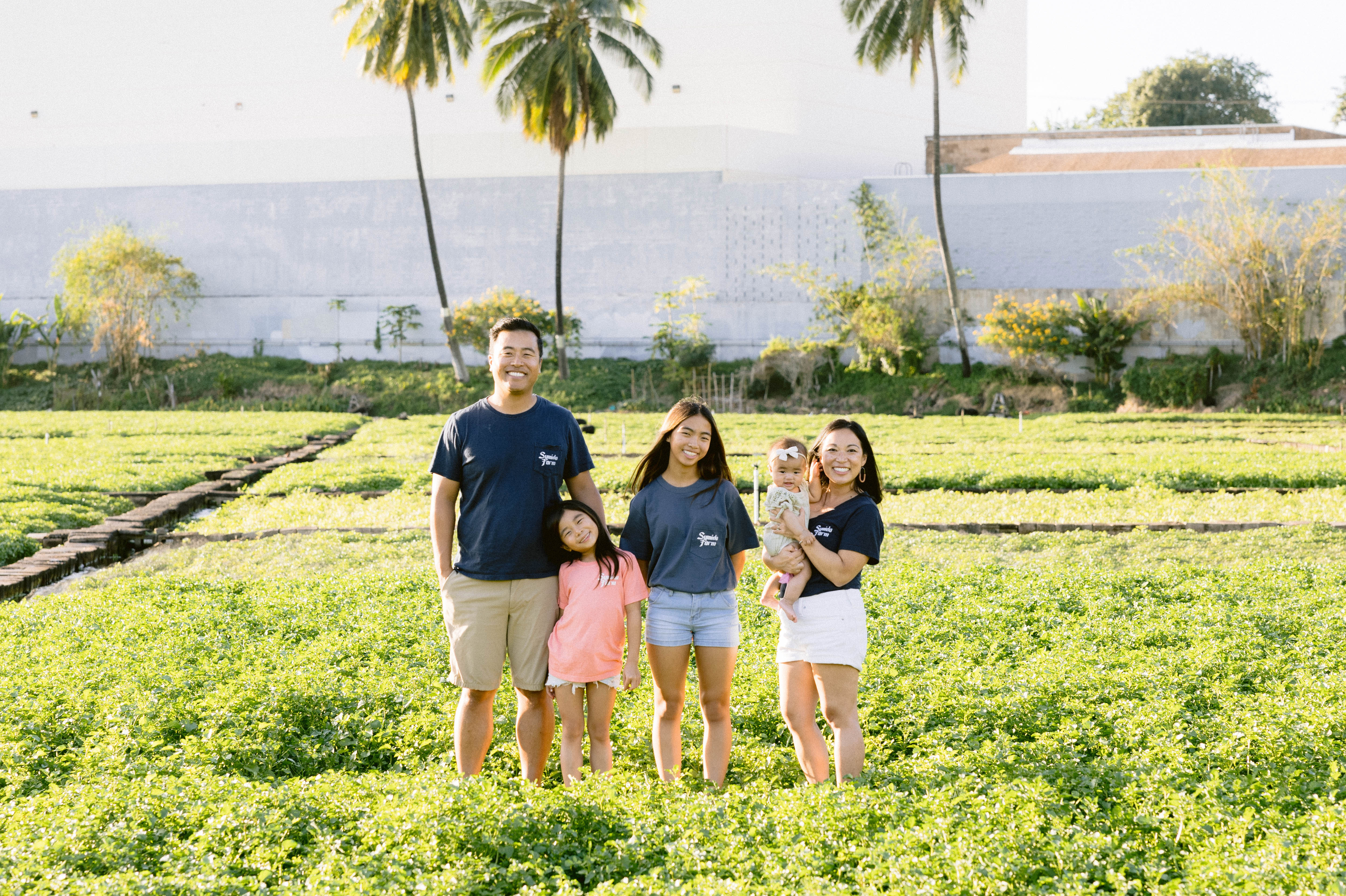 A family of five stands together in a green farm field, with two adults, two children, and a baby posing among rows of crops. Palm trees and garden plots are visible in the background, highlighting a peaceful agricultural setting.