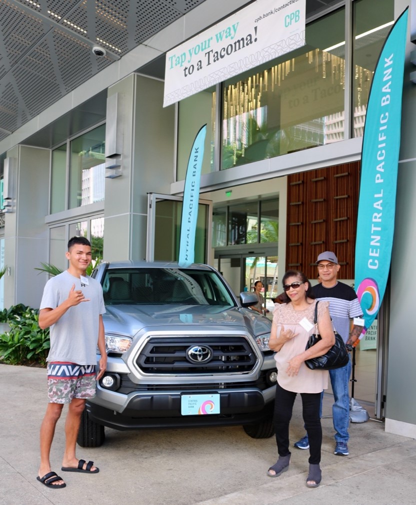 A silver Toyota Tacoma is displayed outside a Central Pacific Bank branch beneath a banner reading “Tap your way to a Tacoma!” Three people pose beside the truck, making shaka gestures during a promotional vehicle giveaway event.