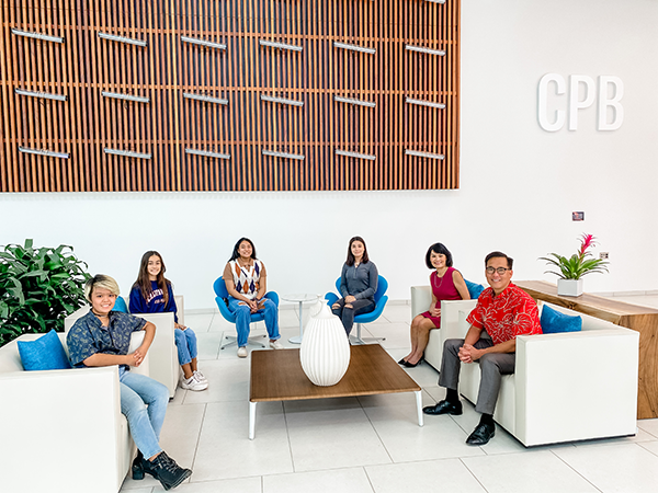 A group of six people sits in a modern Central Pacific Bank lobby, gathered around a coffee table beneath a decorative wooden wall feature and the CPB logo. Bright seating, indoor plants, and an open layout create a welcoming meeting space.