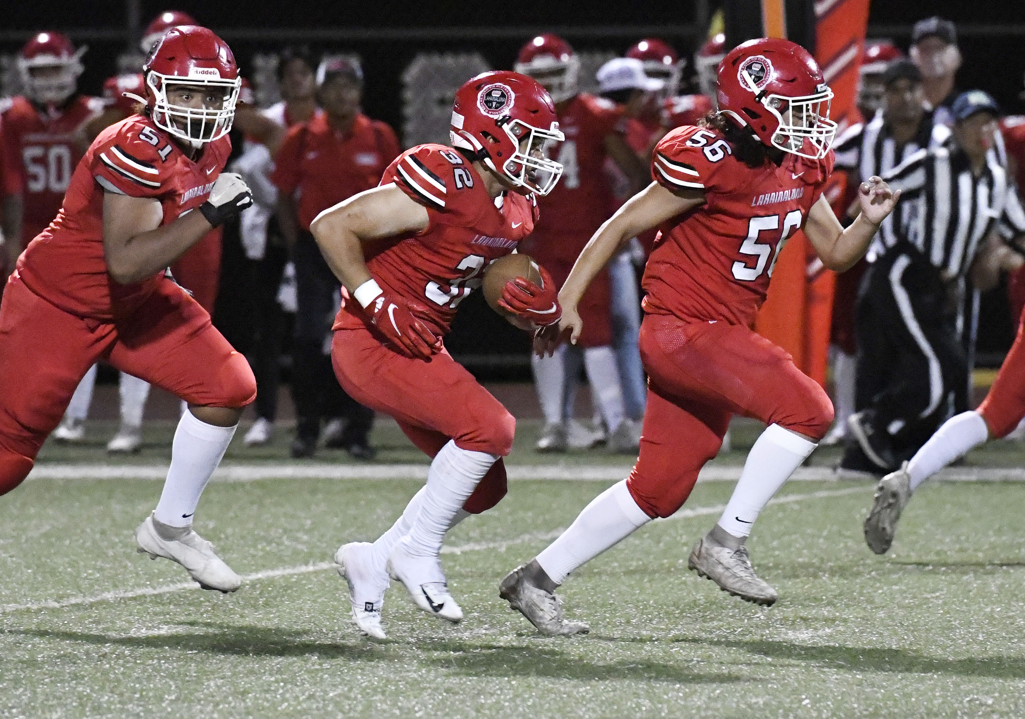 A football player in a red uniform runs with the ball during a nighttime game while teammates in matching red uniforms block and sprint alongside. Coaches, officials, and other players are visible on the sideline in the background.