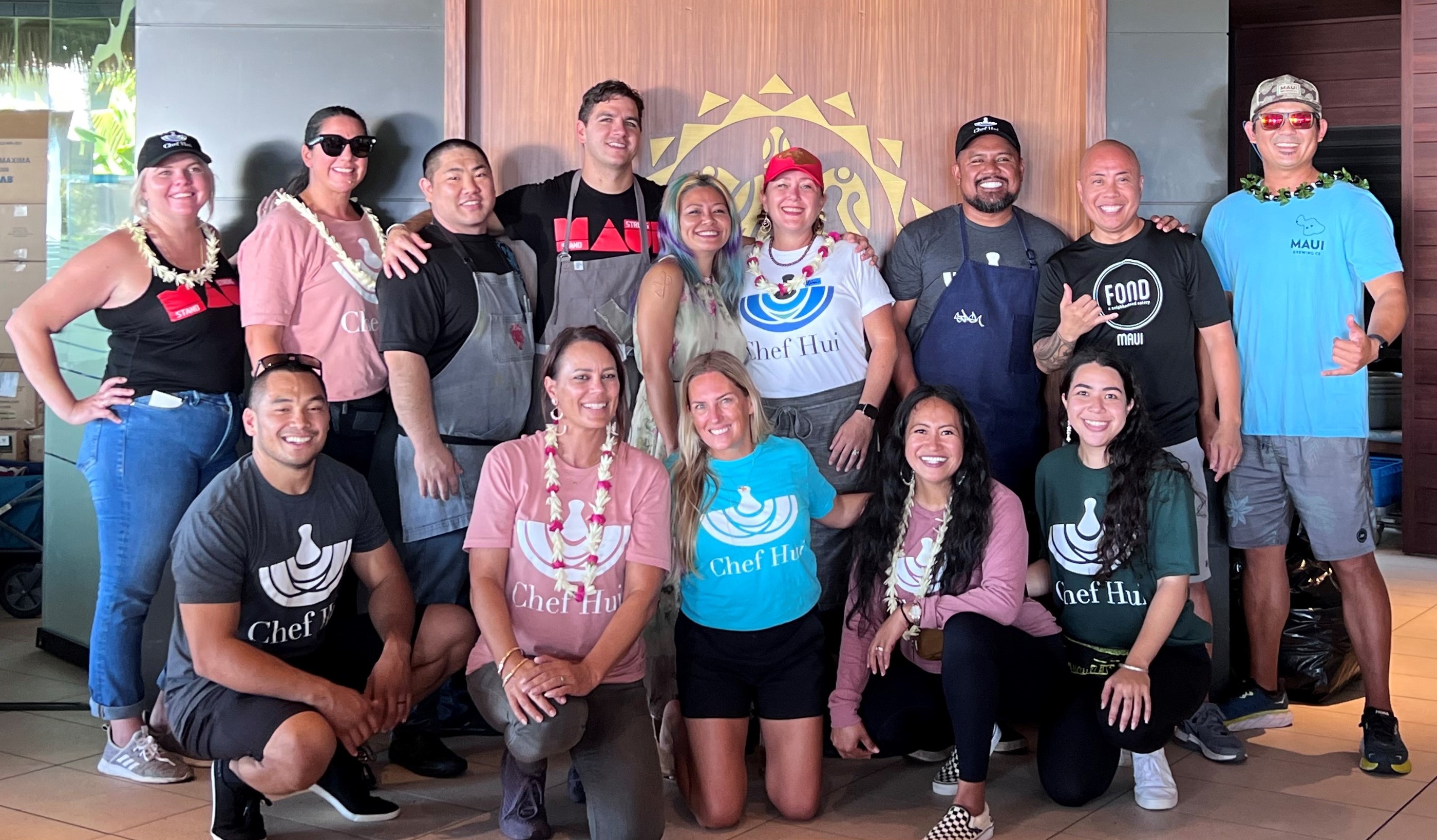 A group of volunteers and community partners pose together indoors, many wearing Chef Hui shirts and flower leis. The group stands in front of a wood-paneled wall, highlighting a collaborative food relief or community service initiative.