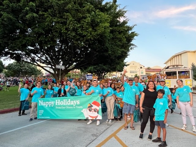 A group of community members wearing matching teal shirts poses along a street holding a “Happy Holidays From Our Ohana to Yours” banner. The festive gathering takes place near a park filled with attendees, with large trees, buildings, and a trolley-style vehicle visible in the background.