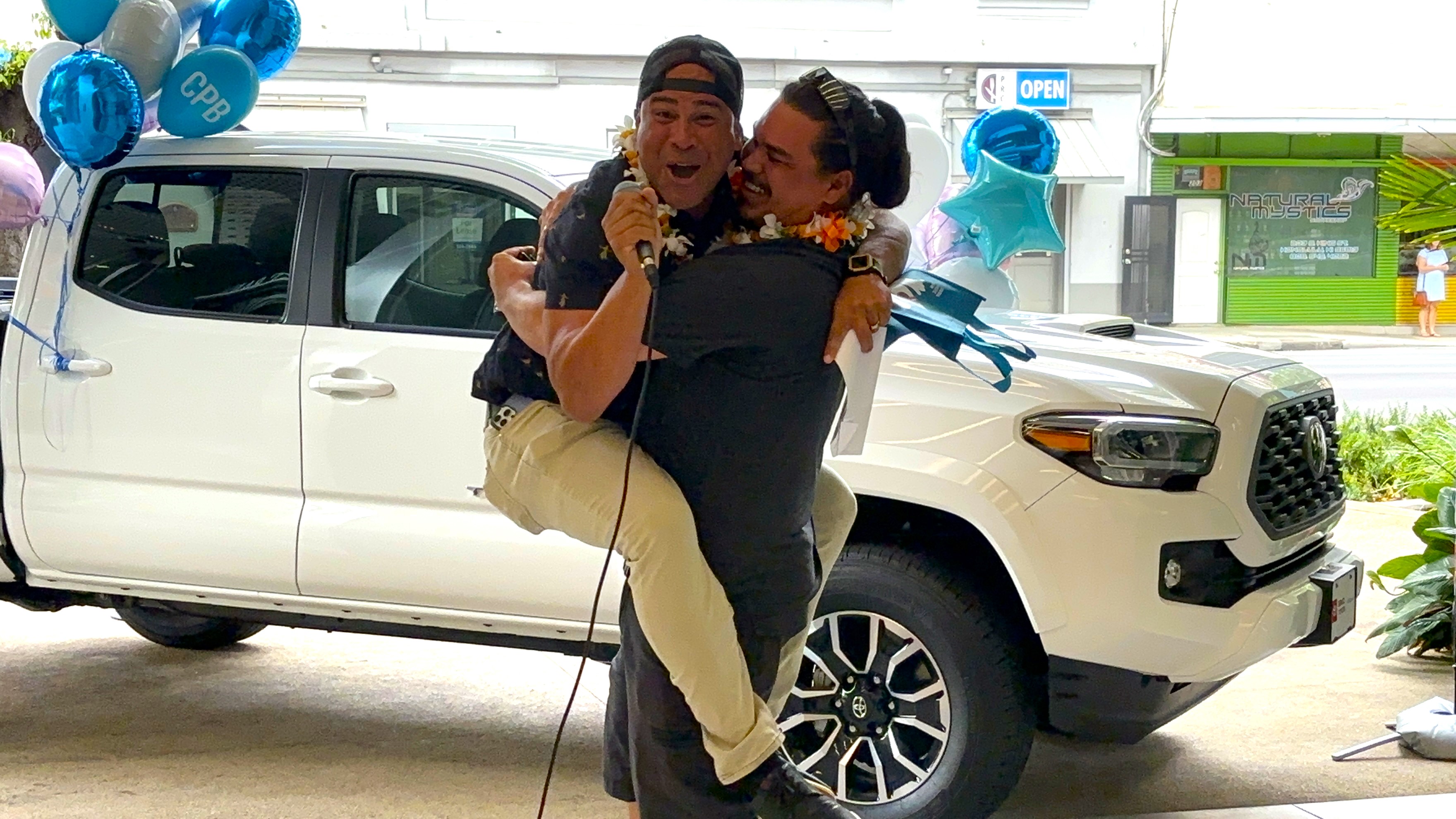 A person holding a microphone is lifted in celebration by another person wearing a flower lei in front of a white Toyota Tacoma decorated with balloons. The scene takes place outside a Central Pacific Bank branch during a truck giveaway event.