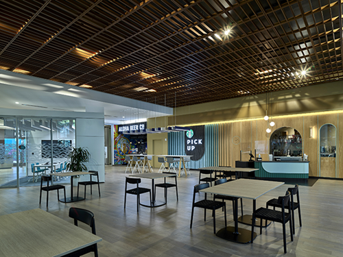 A modern indoor food hall features open seating, wood-accented architecture, and several dining tables arranged throughout the space. Visible storefronts include a pickup counter and food vendors, creating a contemporary communal dining and gathering area.