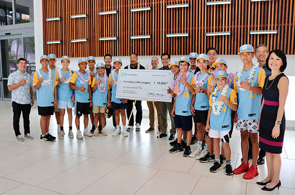 A youth baseball team and community supporters gather in the Central Pacific Bank lobby to present a $10,000 CPB Foundation donation check to the Honolulu Little League. Team members in matching blue and yellow uniforms pose beneath a large decorative wood wall installation while holding the ceremonial check.
