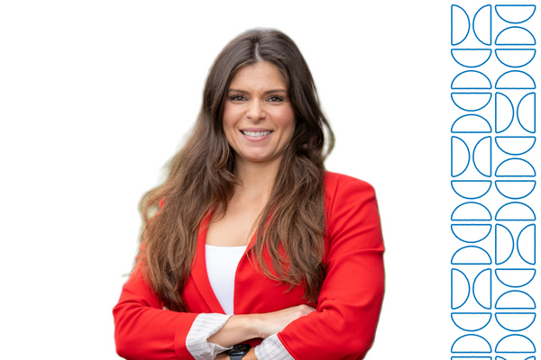 A professional portrait of a person wearing a bright red blazer over a white top, standing with arms crossed against a light gray background. A blue geometric design element appears along the right side of the image.