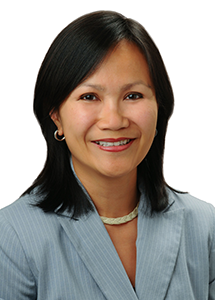 A professional headshot of a person wearing a light blue pinstripe blazer and a necklace, photographed against a light gray background.