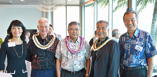A group of five people wearing business attire and floral leis pose together at an indoor networking or community event, with floor-to-ceiling windows and palm trees visible in the background.