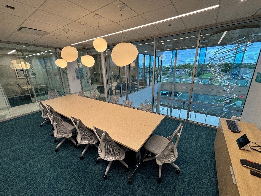 A modern conference room features a large meeting table surrounded by office chairs, with pendant lights hanging overhead and floor-to-ceiling windows overlooking the street outside. Glass-walled offices and contemporary furnishings create a bright, professional workspace.