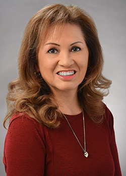 A professional headshot of a person wearing a red long-sleeve top and a pendant necklace, photographed against a gray studio background.