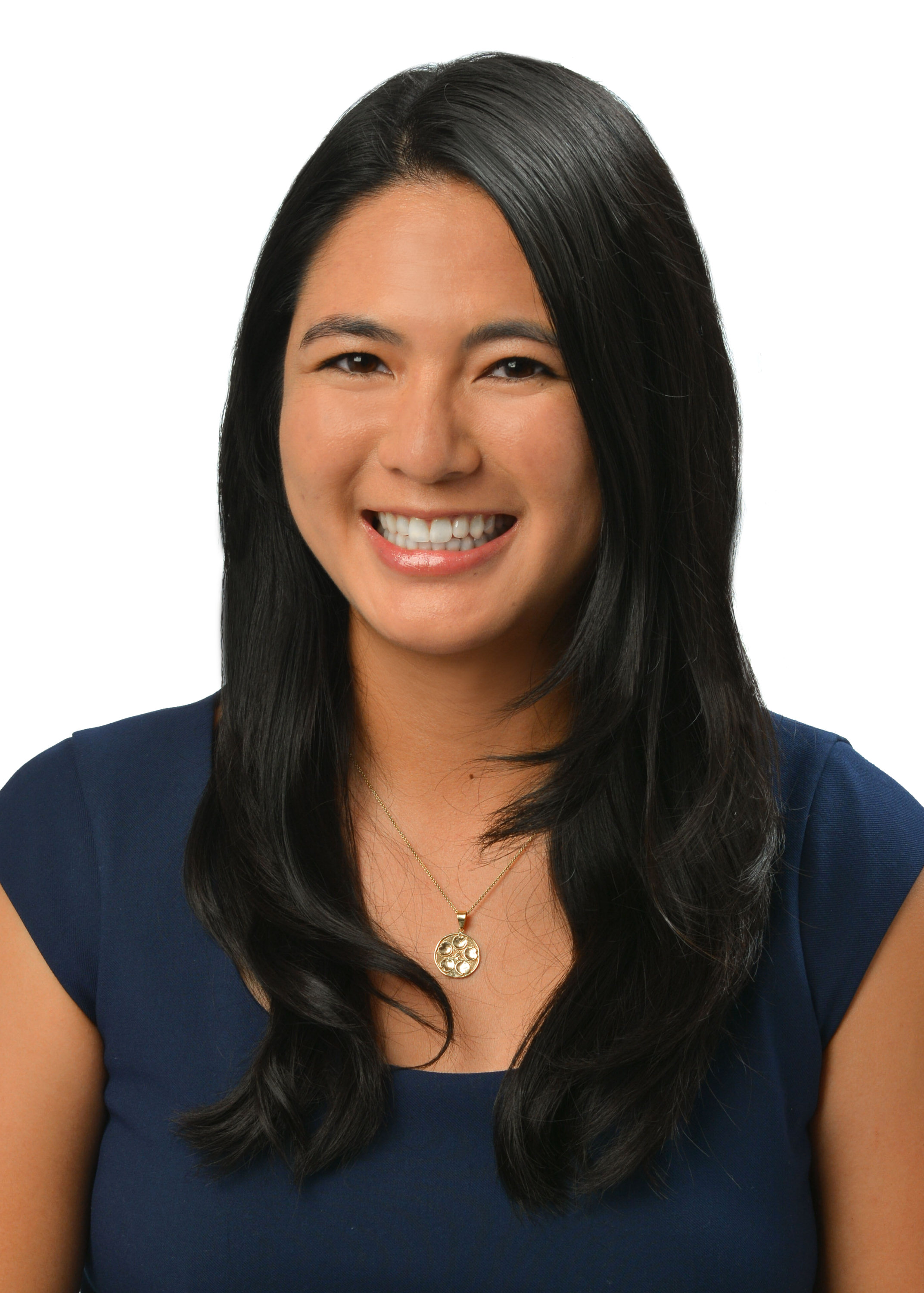 A professional headshot of a person with long dark hair wearing a navy blue top and a gold pendant necklace, photographed against a light gray background.