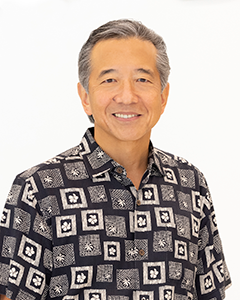 A professional headshot of a person wearing a black-and-white patterned aloha shirt, photographed against a plain white background.