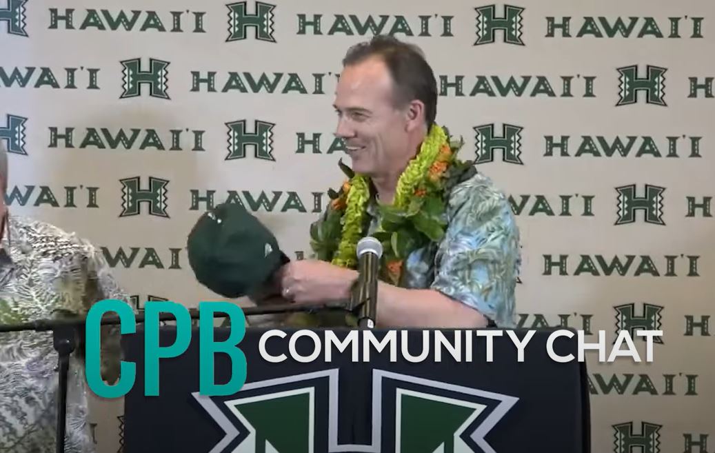 A person wearing a lei stands at a podium during a University of Hawaiʻi event, holding a green cap in front of a backdrop repeating the university’s “H” logo and name. The image includes the text overlay “CPB Community Chat.”