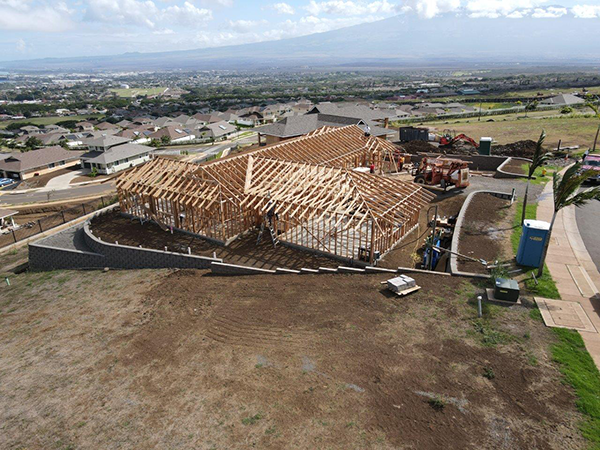 Aerial view of a residential construction project showing a large wood-framed building under construction within a hillside neighborhood. Rows of completed homes, construction equipment, and distant mountain views surround the site.