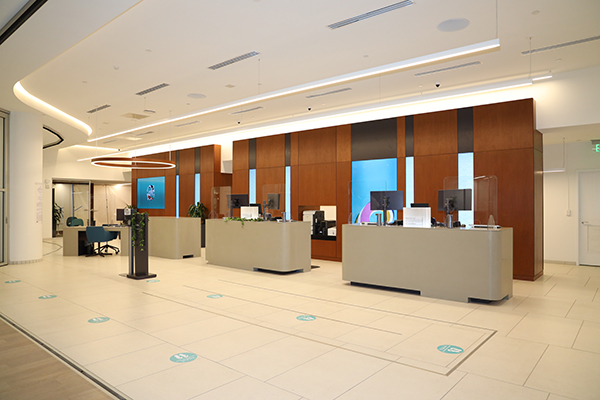 A modern Central Pacific Bank branch features spacious teller stations, wood-paneled accent walls, blue digital displays, and social distancing markers on the floor. The bright, contemporary banking space includes protective screens at service counters and a circular overhead light fixture.