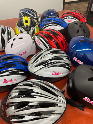 A collection of bicycle helmets in various colors, including white, black, blue, red, and gray, is arranged on a table. Several helmets display the word “Shaka” on the front, highlighting safety gear prepared for a cycling or community program.