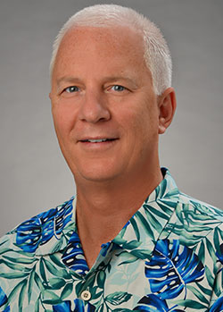 A professional headshot of a person wearing a blue and green floral Hawaiian shirt against a gray studio background.