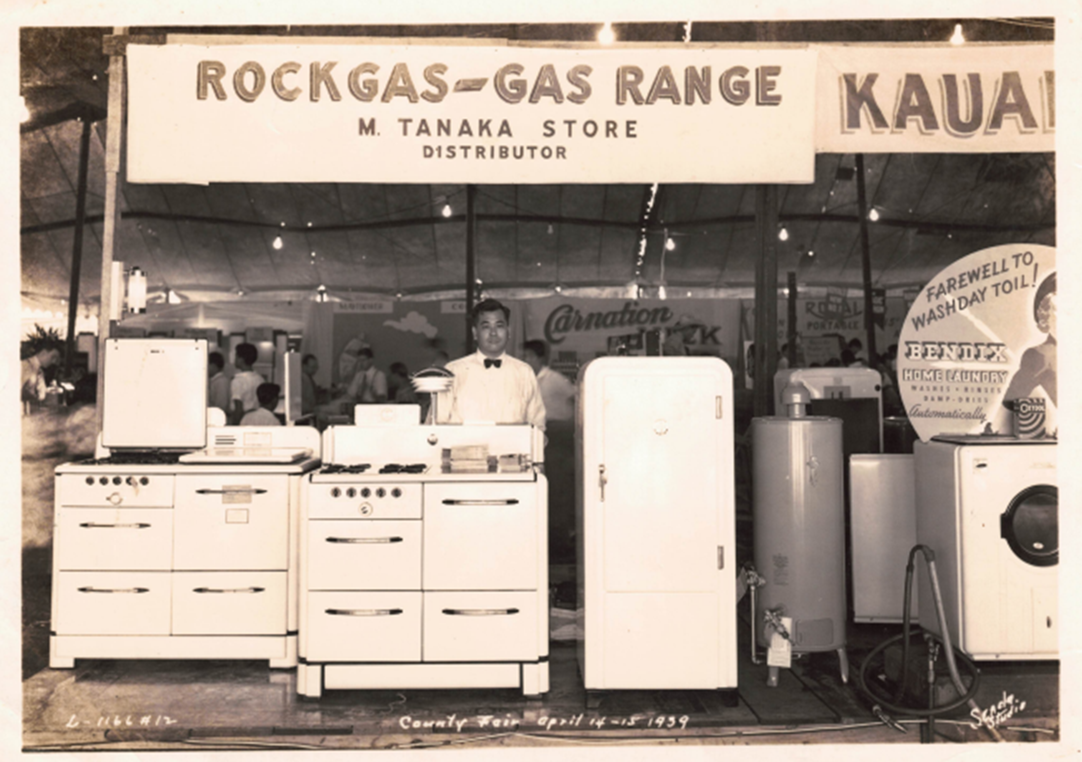 A historic black-and-white photograph shows a home appliance display at a county fair, featuring gas ranges, a refrigerator, a water heater, and a washing machine. A large sign above the booth reads “ROCKGAS-GAS RANGE” with “M. Tanaka Store Distributor,” while fair attendees can be seen in the background.
