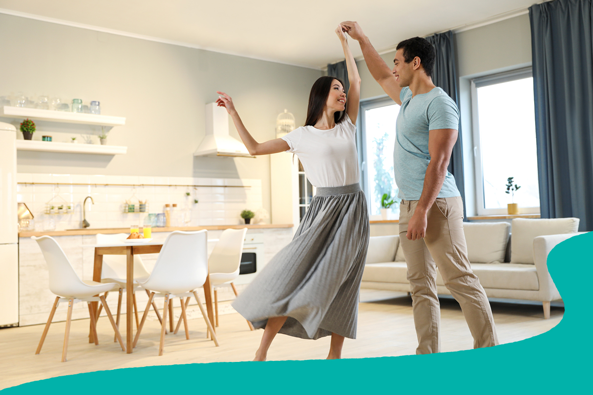 A couple dances together in a bright, modern living room with an open kitchen and dining area in the background. Large windows, light-colored furnishings, and natural light create a warm and welcoming home setting.