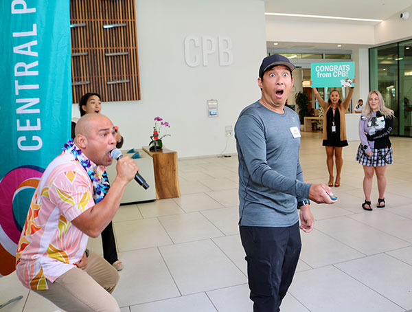 A person stands in the Central Pacific Bank lobby during a surprise celebration while another person speaks into a microphone nearby. A screen in the background displays the message “Congrats from CPB!” as employees look on in the bright, modern lobby.