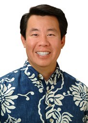 A professional headshot of a person wearing a blue Hawaiian shirt with a white floral pattern, photographed against a light gray background.