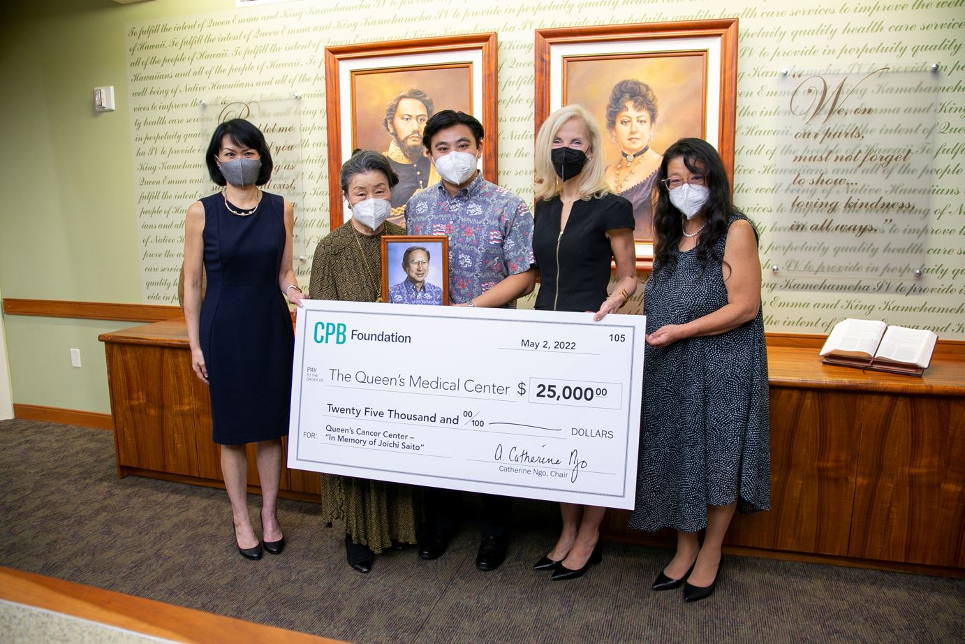 Five people wearing face masks pose indoors while holding a large CPB Foundation donation check for $25,000 benefiting The Queen’s Medical Center. The group stands beneath framed portraits and displays a memorial photo during the check presentation ceremony.