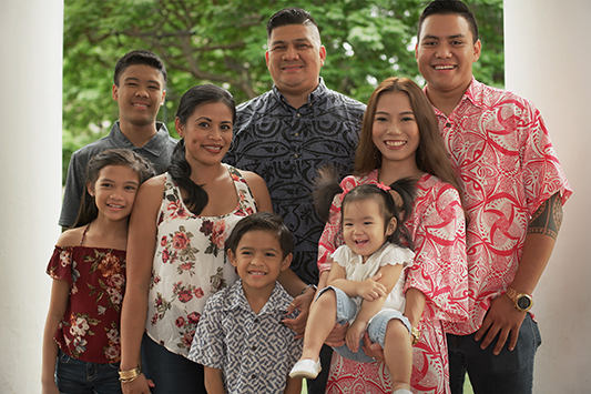 A multigenerational family portrait showing eight people posing together outdoors between white columns, with green trees in the background. Several individuals wear floral-patterned clothing, and a young child is being held in the front row.