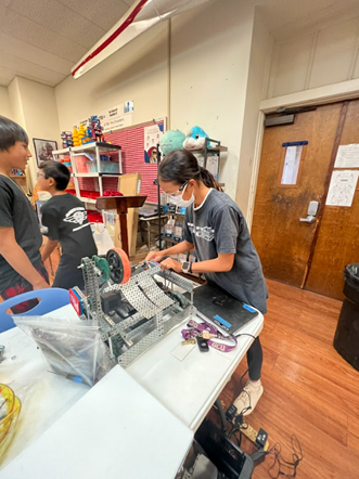 A student works on assembling a robotics project at a table in a classroom, using tools and mechanical parts while other students collaborate nearby. Shelves with supplies, posters, and robotics equipment are visible in the background.