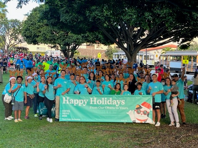A large group of community members wearing matching teal shirts gathers in a park behind a banner that reads, “Happy Holidays From Our Ohana to Yours.” The Central Pacific Bank-sponsored event takes place under large shade trees, with families and additional participants visible throughout the background.
