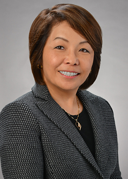 A professional headshot of a person wearing a textured gray blazer over a black top, photographed against a gray studio background.
