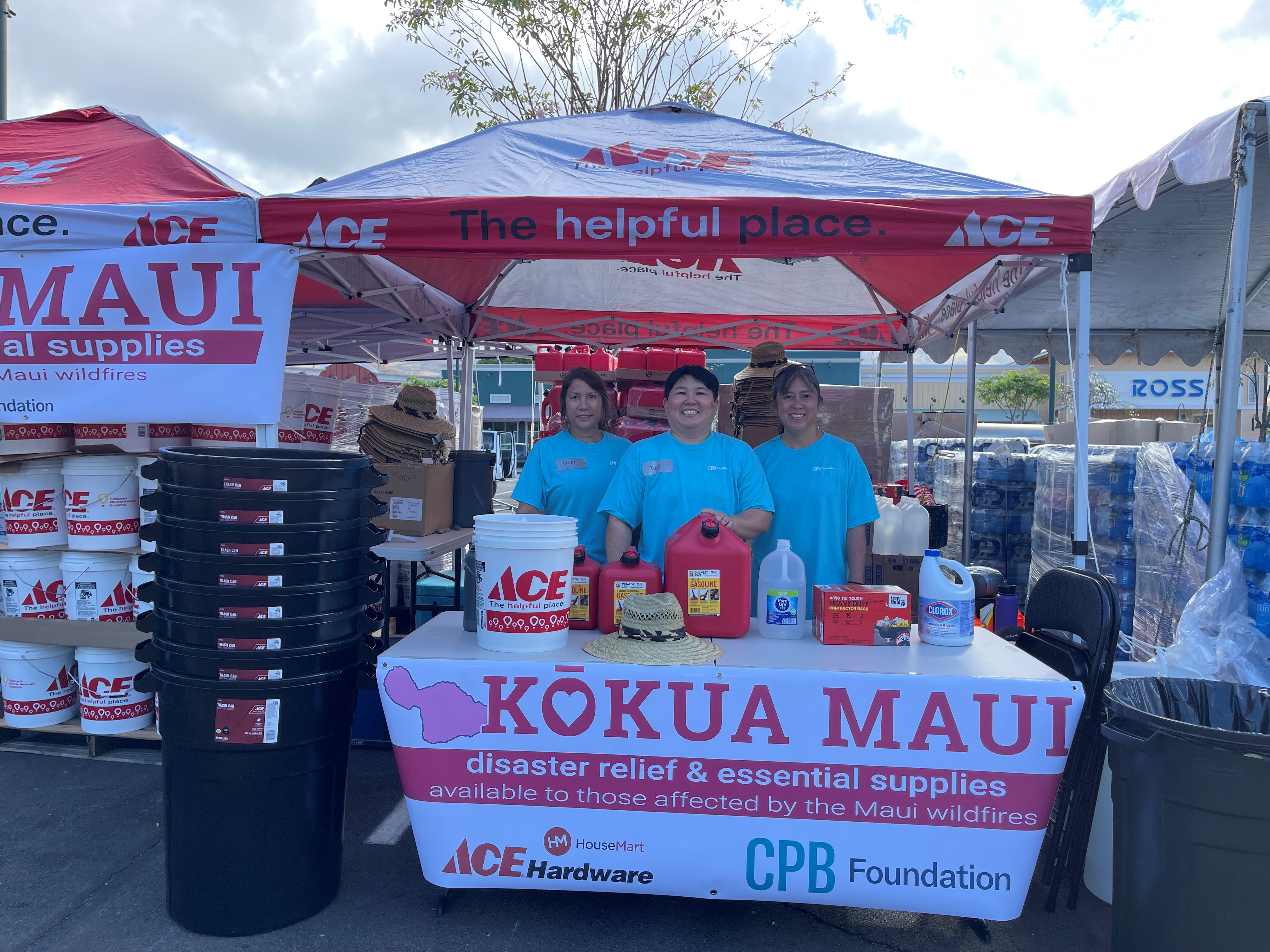 Three volunteers stand behind a Kōkua Maui disaster relief booth stocked with fuel containers, cleaning supplies, water, and emergency kits. The booth, supported by ACE Hardware and the CPB Foundation, provides essential supplies for individuals affected by the Maui wildfires.