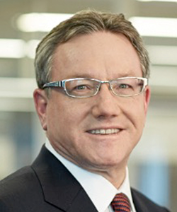 A professional headshot of a person wearing glasses, a dark business suit, white shirt, and red striped tie, photographed against a softly blurred office background.