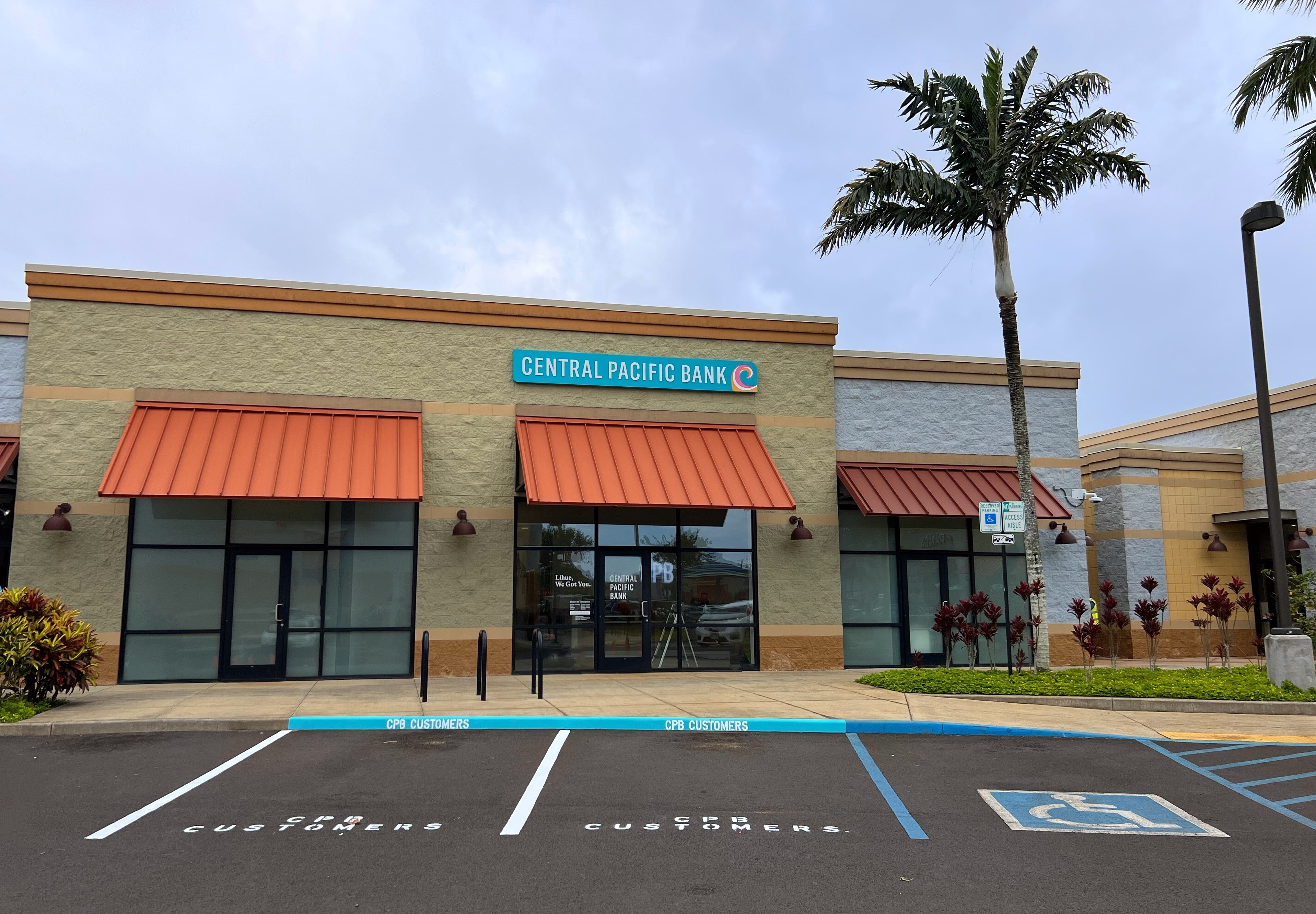 A Central Pacific Bank branch with orange awnings and prominent exterior signage is shown from the parking lot, with palm trees and landscaped greenery surrounding the building. Designated customer parking spaces and an accessible parking area are visible in front of the entrance.