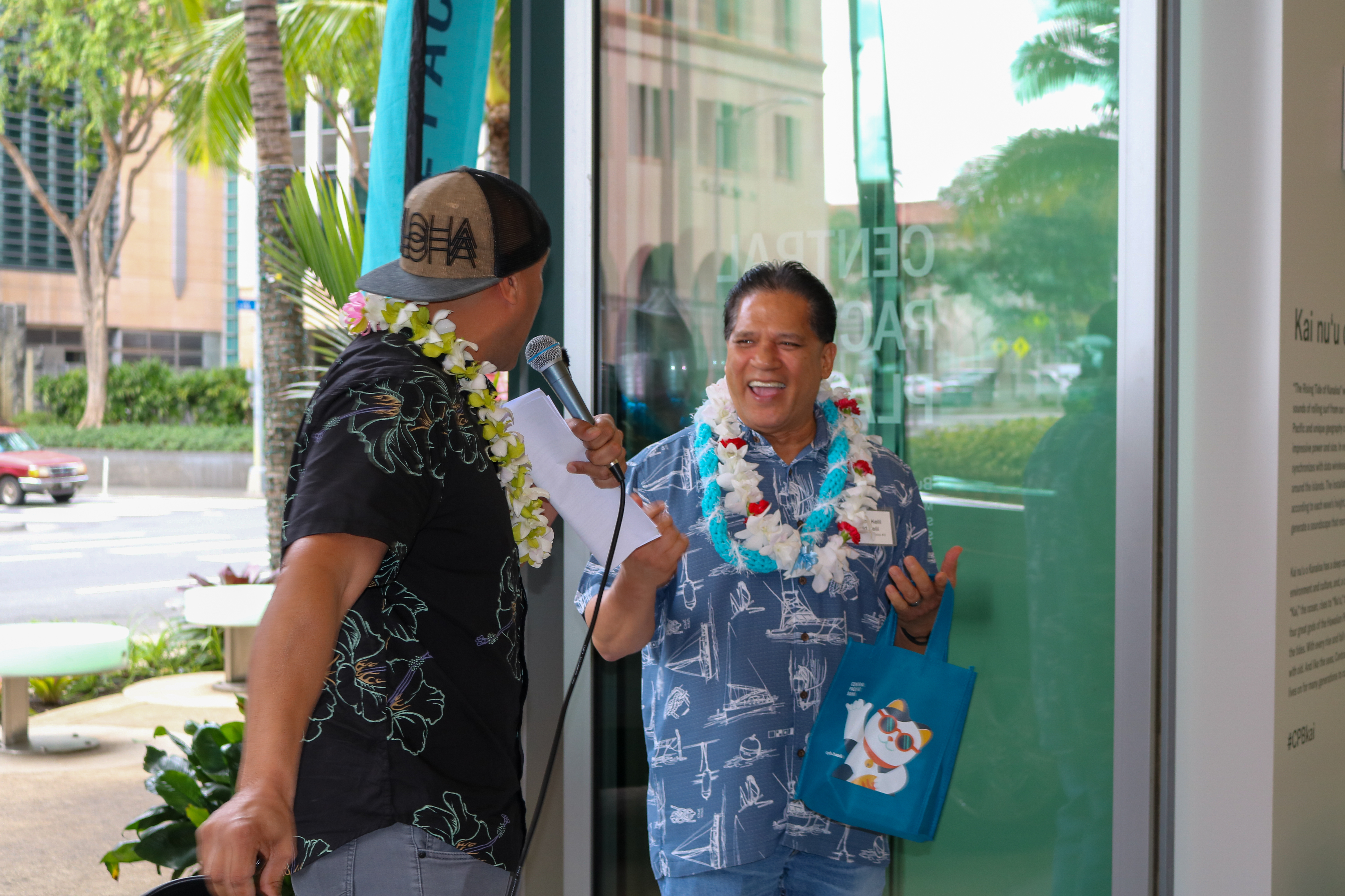 A person wearing a flower lei holds a gift bag while speaking with an event host holding a microphone inside a Central Pacific Bank branch. The pair stand near large windows during a promotional celebration or prize giveaway event.