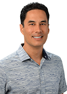 A professional headshot of a person wearing a light blue patterned collared shirt, photographed against a light gray background.