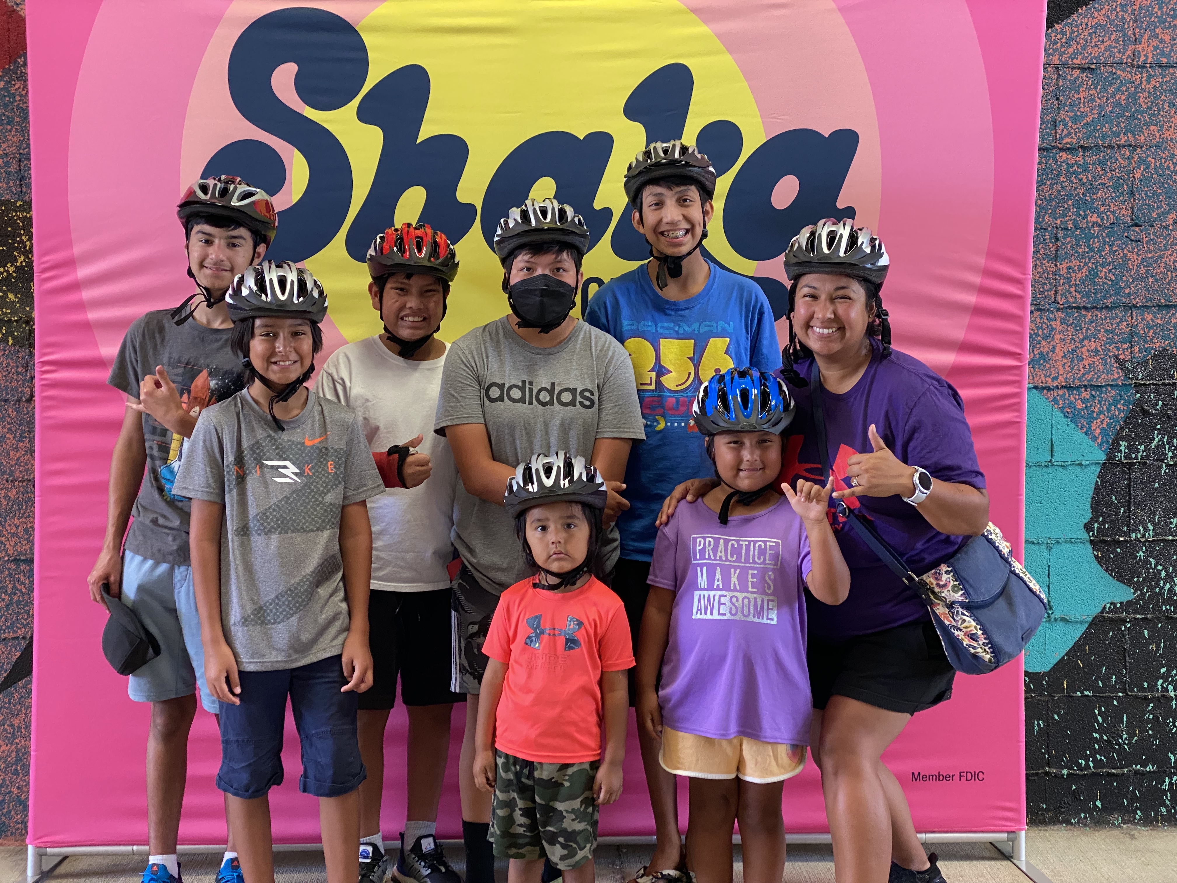 A group of children and adults wearing bicycle helmets pose in front of a bright pink “Shaka” backdrop. Several participants make shaka hand gestures, highlighting a community cycling or helmet safety event.