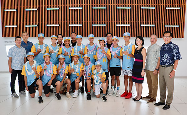 A youth baseball team in matching blue and yellow uniforms poses for a group photo in the Central Pacific Bank lobby with community supporters. Several team members wear leis and make shaka gestures while standing beneath a large decorative wood wall installation.