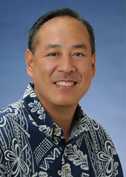 A professional headshot of a person wearing a blue patterned Hawaiian shirt, photographed against a blue studio background.