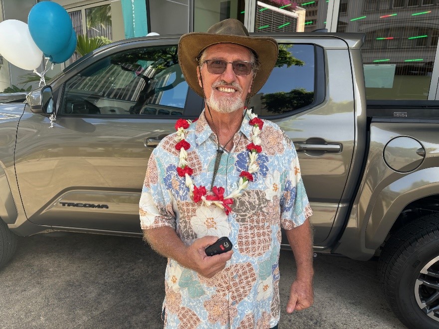 A person wearing a floral shirt, lei, sunglasses, and a wide-brimmed hat stands in front of a Toyota Tacoma, holding a vehicle key fob. Balloons and the pickup truck are visible in the background, suggesting a vehicle giveaway or award presentation event.