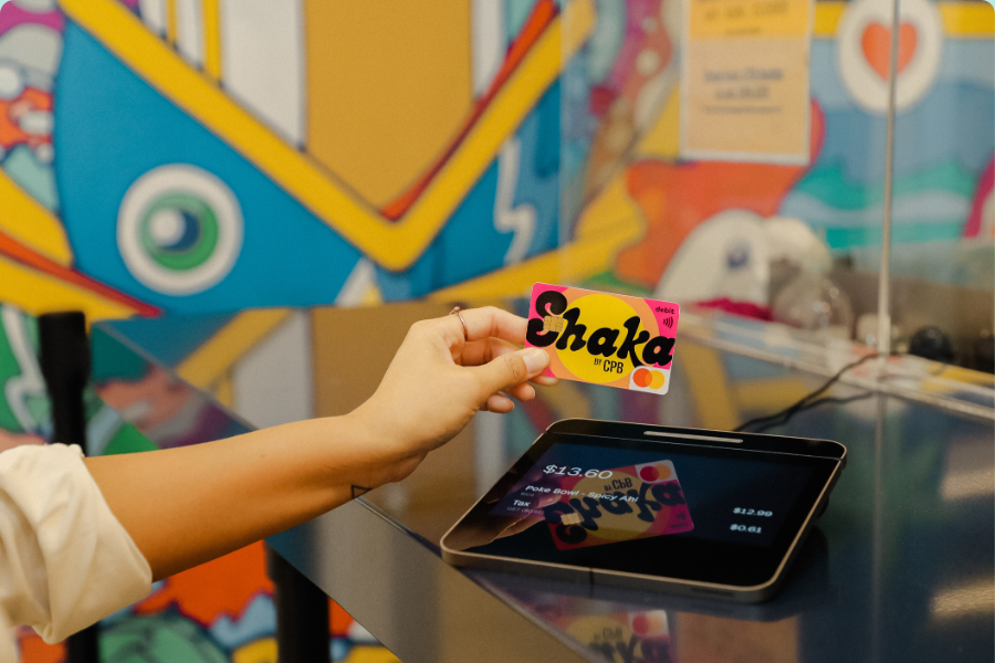 Person holding a CPB Shaka debit card over a payment terminal to complete a contactless payment at a checkout counter.
