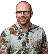 A professional headshot of a person wearing glasses and a light-colored Hawaiian shirt with a floral pattern, photographed against a light gray background.